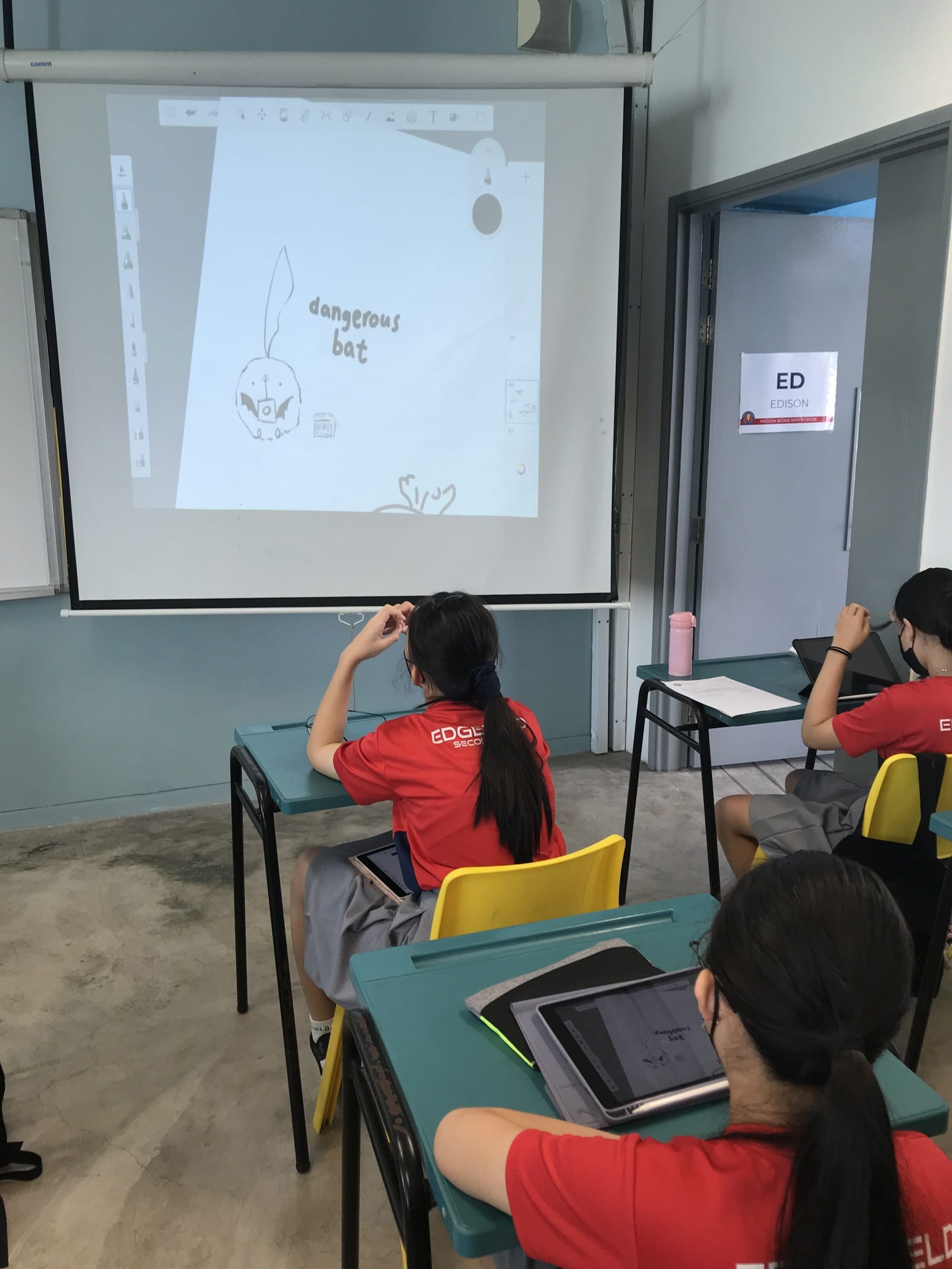 Students in red uniforms sit at desks with tablets, facing a projected screen showing a drawing labeled dangerous bat.
